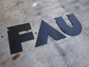 auf einem rostigen Metalldach liegen drei ausgesägte Holzbuchstaben: FAU